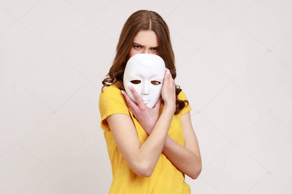 Portrait of teenager girl with brown hair in yellow casual style T-shirt covering half of face with white mask, multiple personality disorder. Indoor studio shot isolated on gray background.
