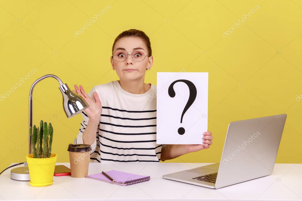 Smart woman office employee sitting on workplace and holding question mark, having doubtful expression, don't know the solution, Indoor studio studio shot isolated on yellow background.