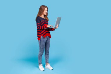 Süveter giymiş, elinde taşınabilir bilgisayarla dizüstü bilgisayara gülümseyen ve işten memnun bir şekilde bakan çekici bir kadının tam boy portresi. Mavi arka planda kapalı stüdyo çekimi.