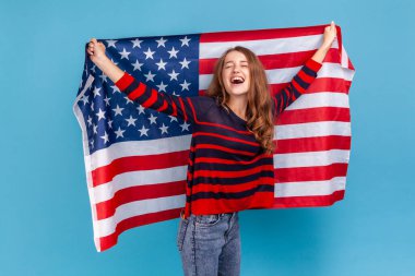 Çizgili bir süveter giyen, elinde kocaman bir Amerikan bayrağı tutan ve mutlu bir şekilde bağıran heyecanlı genç bir kadın ulusal bayramı kutluyor. Mavi arka planda kapalı stüdyo çekimi.