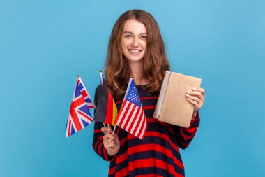 Çizgili sıradan süveter giyen, elinde Almanca, ABD ve Uk 'un kitap bayrakları tutan yabancı dil öğrenen, yurtdışında eğitim alan pozitif öğrenci kız. Mavi arka planda kapalı stüdyo çekimi.