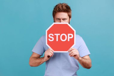 Ciddi derecede sert sakallı bir adamın portresi yüzünün yarısını kırmızı trafik levhasıyla kaplıyor, uyarı, içki yasağı konsepti, otoriter ifade. Mavi arka planda kapalı stüdyo çekimi.