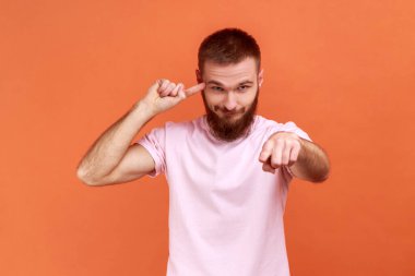 Portrait of bearded man showing stupid gesture and pointing to camera, blaming for insane plan, crazy idea, dumb suggestion, wearing pink T-shirt. Indoor studio shot isolated on orange background.