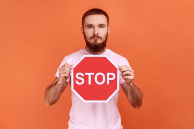 Elinde kırmızı dur tabelası olan ciddi bir sakallı adamın portresi ciddi bir ifadeyle kameraya bakıyor, pembe tişört giyiyor. Turuncu arka planda kapalı stüdyo çekimleri.