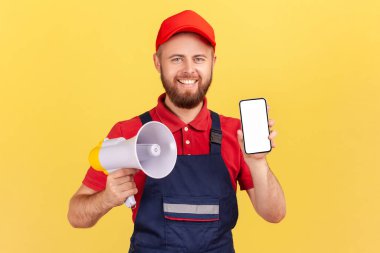 Mavi tulumlu ve kırmızı şapkalı mutlu bir işçi duyuru yapmak için megafon ve reklam için boş ekranlı cep telefonu tutuyor. Sarı arka planda kapalı stüdyo çekimleri.
