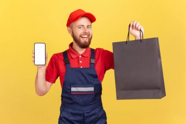 Üniformalı mutlu teslimatçı elinde alışveriş torbalarıyla akıllı telefonu boş vitrinle gösteriyor, mağazadan hızlı teslimat, servis endüstrisi. Sarı arka planda kapalı stüdyo çekimleri.