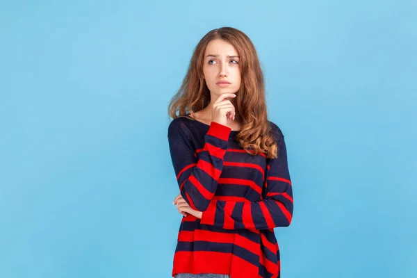 戦略について考えて あごを保持立って縞模様のカジュアルなスタイルのセーターを身に着けている若い女性は 離れて見て 解決策を見つける 青い背景に隔離された屋内スタジオショット — ストック写真