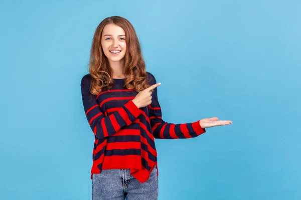 Good looking woman wearing striped casual style sweater, presenting copy space on her palm, showing empty place for commercial text or goods. Indoor studio shot isolated on blue background.