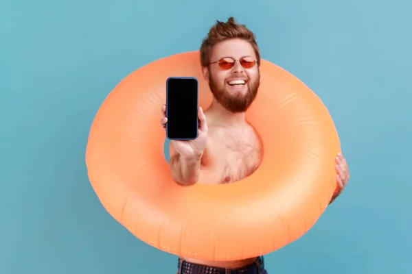 Portrait of smiling positive man holding orange rubber ring and showing phone with blank screen for advertisement, looking at camera with toothy smile. Indoor studio shot isolated on blue background.