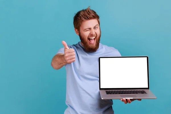 正面胡子男子拿着笔记本电脑的肖像 屏幕空旷 对电脑应用感到满意 看着相机 露出大拇指 室内拍摄被蓝色背景隔离 — 图库照片