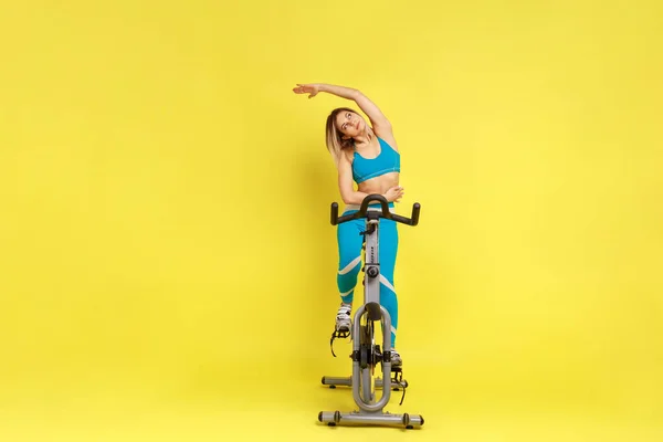 ジムマシンの自転車で働く女性の完全な長さの肖像画と彼女の腕 青いスポーツウェアを着てストレッチ 黄色の背景に隔離された屋内スタジオショット — ストック写真