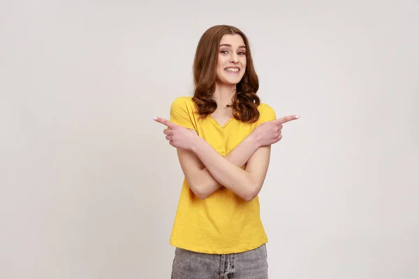 黄色のTシャツに身を包んだ若い年齢の正の非常に幸せな女性の肖像画は 手を交差し 両側にスペースをコピーすることを指して立っている 灰色の背景に隔離された屋内スタジオショット — ストック写真