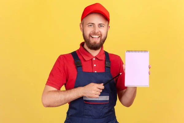 青い全体と赤いキャップを身に着けている楽観的な笑顔の労働者は 快適な笑顔で紙のノートブックを指して コピースペースを提示します 黄色の背景に隔離された屋内スタジオショット — ストック写真