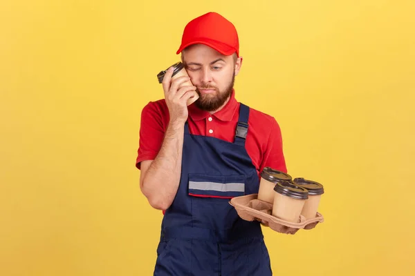 疲れた疲れ労働者は 閉じた目で立って 昼寝をし コーヒーの使い捨てカップを保持し 全体とキャップを身に着けているハードワークの後に眠く感じる 黄色の背景に隔離された屋内スタジオショット — ストック写真