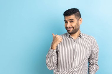 Hoş görünümlü sakallı bir adamın portresi. Parmağını fotokopi odasına doğru uzatıyor. Reklam için bölgeyi gösteriyor, çizgili gömlek giyiyor. Mavi arka planda kapalı stüdyo çekimi.