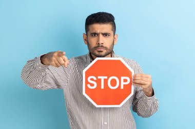 Ciddi bir iş portresi büyük kırmızı yol tabelasını tutuyor ve seni işaret ediyor, kameraya bakıyor, çizgili gömlek giyiyor. Mavi arka planda kapalı stüdyo çekimi.