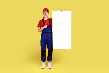 Reklam ya da tanıtım metni için boş yeri olan beyaz kağıt pankartı tutan ve işaret eden tam boy kadın portresi. Sarı arka planda kapalı stüdyo çekimleri.