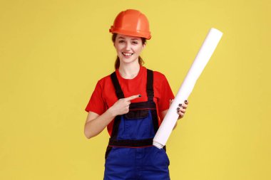 Tatmin olmuş kadın mimarın tasviri planlara işaret ediyor, kameraya gülümseyerek bakıyor, tulum giyiyor ve koruyucu başlık takıyor. Sarı arka planda kapalı stüdyo çekimleri.