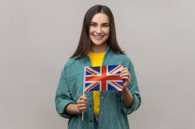İngiliz bayrağını tutan mutlu kadının portresi, dişlek gülüşlü kameraya bakıyor, vatansever, günlük tarz ceket giyiyor. Gri arka planda izole edilmiş kapalı stüdyo görüntüleri.