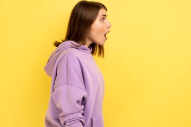 Şok olmuş bir ifadeye sahip, ağzı açık, mor kapüşonlu bir kadın profili. Sarı arka planda kapalı stüdyo çekimleri.