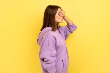 Bakmak istemiyorum. Yandan bakan kadın portresi. Gözlerini ayırmayan ve mor kapüşonlu, utanç verici, iğrenç bir şeye bakmaktan kaçınan kadın portresi. Sarı arka planda kapalı stüdyo çekimleri.