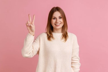 Zafer ya da barış jesti gösteren, geniş gülümsemeli, şanslı kazanan, gelecekteki başarısı hakkında iyimser hisseden, beyaz kazak giyen bir kadın. Pembe arka planda kapalı stüdyo çekimleri.