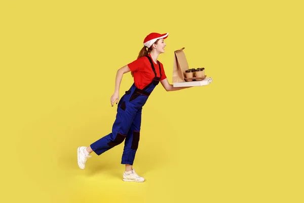 Side view portrait of delivery woman being hurry to deliver coffee with pizza, fast delivery service, wearing overalls and red cap. Indoor studio shot isolated on yellow background.
