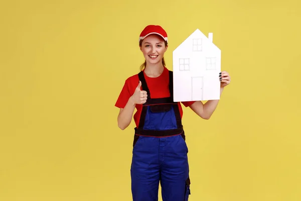 Handy woman holding paper house and showing thumb up, recommend home repair services and maintenance, wearing overalls and red cap. Indoor studio shot isolated on yellow background.