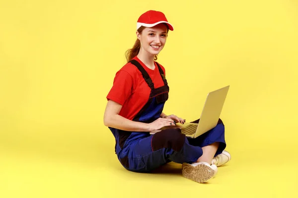 Side view portrait of satisfied handy woman sitting on floor with crossed legs and working on laptop, looking at camera, wearing overalls and red cap. Indoor studio shot isolated on yellow background.