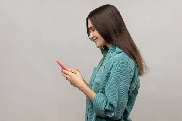 スマートフォンや笑顔でメッセージを読む幸せな女性のサイドビュー 通信のためのモバイルデバイスを使用して ウェブを閲覧 カジュアルスタイルのジャケットを身に着けています 灰色の背景に隔離された屋内スタジオショット — ストック写真