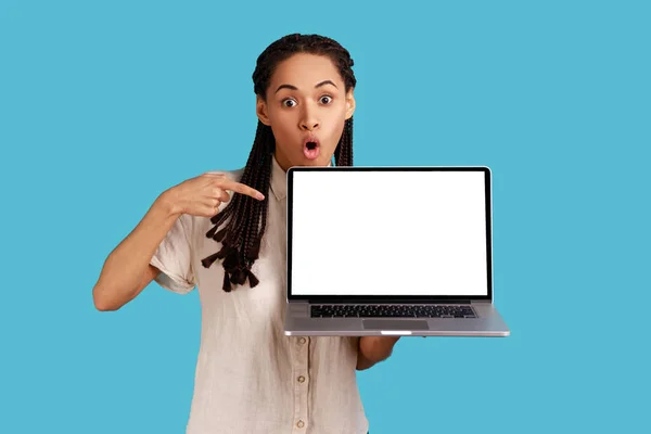 ドレッドロックを持つ印象的な怖がっている女性の関心とスリルから口を開き 空の画面でノートパソコンのポイントは 白いシャツを着て 魅了を感じる 青い背景に隔離された屋内スタジオショット — ストック写真