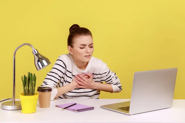 Fatigued ill woman manager sitting at workplace with laptop and holding painful chest, suffering heart attack, myocardial infarction. Indoor studio studio shot isolated on yellow background.