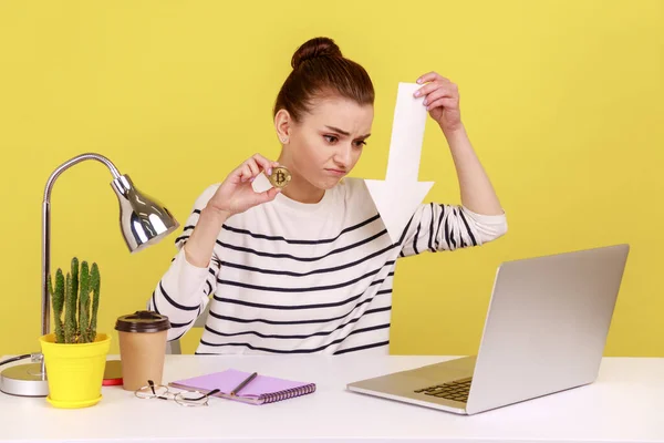 不满意的女人坐在工作场所展示黄金比特币和箭头指向笔记本电脑显示 降低电子货币的等级 在黄色背景下隔离的室内工作室拍摄 — 图库照片