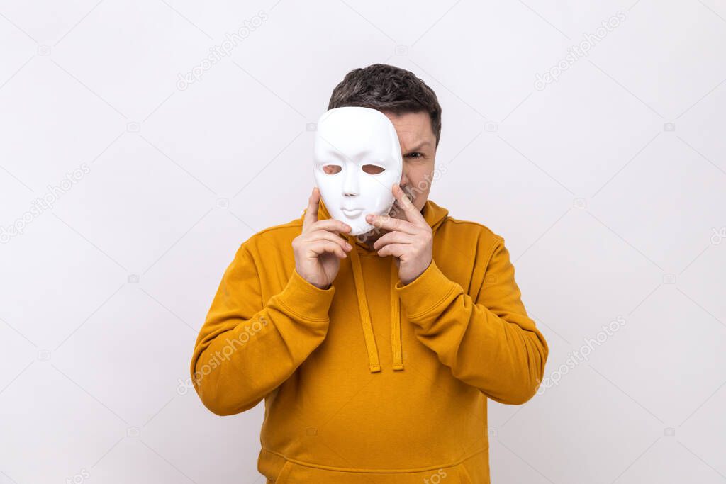 Middle aged man holding white mask, covering face, standing with serious expression, multiple personality disorder, wearing urban style hoodie. Indoor studio shot isolated on white background.