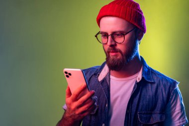Ciddi iddialı hipster bir adamın portresi akıllı telefonda yazı yazması, bahis oynaması, online uygulama kullanması, habercilerle sohbet etmesi. Renkli neon ışıklı arka planda kapalı stüdyo çekimleri.