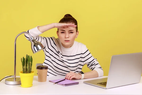 一个年轻女性员工的画像 她带着笔记本电脑坐在工作场所 手放在眼睛上方遥望远方 目不转睛地搜寻 在黄色背景下隔离的室内工作室拍摄 — 图库照片