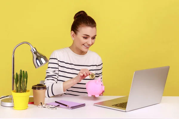 Portrait of happy smiling dark haired woman investing in bitcoins, putting golden crypto coin into piggy bank and looking at laptop display. Indoor studio studio shot isolated on yellow background.