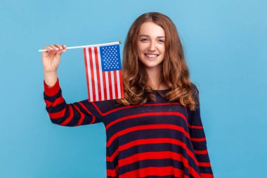 Güzel bir kadın çizgili bir süveter giyiyor, Amerika Birleşik Devletleri bayrağı sallıyor, ulusal bayramı, vatanseverliği, bağımsızlığı kutluyor. Mavi arka planda kapalı stüdyo çekimi.