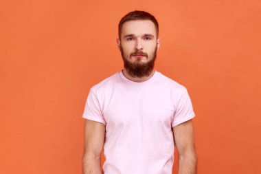Kendine güvenen sakallı bir adamın portresi, ciddi bir ifadeyle kameraya bakıyor, gülümseyen kararlı bir adam, pembe tişört giyiyor. Turuncu arka planda kapalı stüdyo çekimleri.