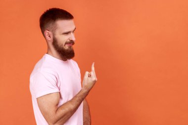 Saldırgan, sinirli, sakallı bir adamın orta parmak gösteren portresi, pembe tişört giyen kaba bir saygısızlık hareketi. Turuncu arka planda kapalı stüdyo çekimleri.