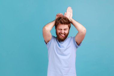 Komik, neşeli, sakallı bir adamın portresi tavşan kulakları ve dili gösteriyor, komik ve heyecanlı görünüyor, oynaşıyor. Mavi arka planda kapalı stüdyo çekimi.