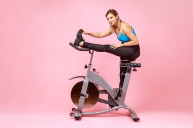 Sıska atletik bir kadının egzersiz bisikletinde otururken esneme hareketleri, antrenmandan önce ısınma hareketleri, spor tayt ve bluz giymesi. Pembe arka planda kapalı stüdyo çekimleri.