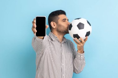 Futbol topunu öpen sakallı yakışıklı bir adamın portresi ve cep telefonunu boş siyah ekranla gösteren, çizgili tişörtlü. Mavi arka planda kapalı stüdyo çekimi.