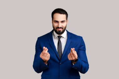 Yakışıklı sakallı işadamının portresi sinsice gülümsüyor ve para jesti yapıyor, ödeme istiyor, harçlık istiyor, resmi tarz takım elbise giyiyor. Gri arka planda izole edilmiş kapalı stüdyo görüntüleri.