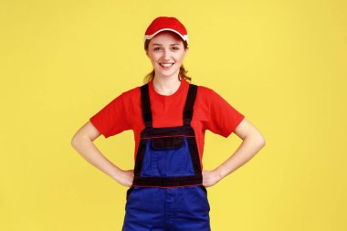 Mutlu işçi kadın portresi, elleri kalçasında, kameraya pozitif özgüvenli bir ifadeyle bakıyor, tulum giyiyor ve kırmızı şapka takıyor. Sarı arka planda kapalı stüdyo çekimleri.