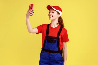 Ellerinde cep telefonuyla selfie çeken ya da canlı yayın desteği alan tatmin olmuş kadın portresi. Sarı arka planda kapalı stüdyo çekimleri.