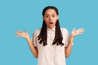 Siyah rastalı kızgın kadın kızgın ifadesini şaşırttı, kollarını kaldırdı, büyük gözleriyle kameraya baktı, beyaz gömlek giydi. Mavi arka planda kapalı stüdyo çekimi.