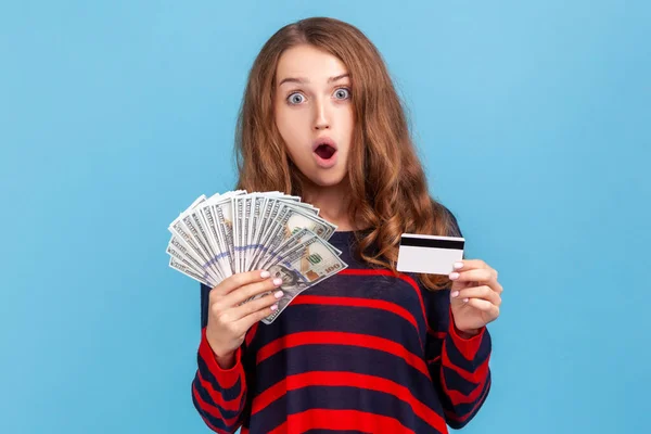 縞模様のカジュアルなスタイルのセーターを着てショックを受けた女性は クレジットカードを保持し 銀行券のファン キャッシュバック 通貨交換に驚いている 青い背景に隔離された屋内スタジオショット — ストック写真
