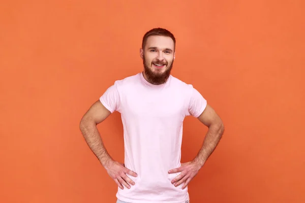 大胡子男人的画像 手托着臀部站在那里 看着相机 表达积极的情绪 心情很好 穿着粉色T恤 在橙色背景下被隔离的室内拍摄 — 图库照片