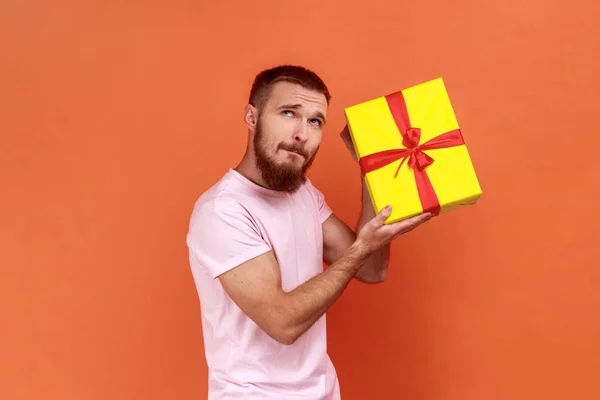 一个好奇的留着胡子的男人站在那里 拿着黄色礼品盒 带着沉思的表情望着别处 心里在想什么 穿着粉红色的T恤 在橙色背景下被隔离的室内拍摄 — 图库照片